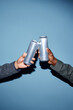 © Seventyfour - Close-up of two people's hands holding and clinking aluminum cans, set against a blue background, signifying a celebratory or friendly moment