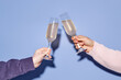 © Seventyfour - Two hands clinking champagne glasses in joyful celebration against a blue backdrop. Clear bubbles ascending inside the glasses, capturing festive spirit