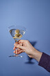 © Seventyfour - Close-up of a hand holding martini glass with three olives against blue background creating an elegant and stylish focus on the drink held