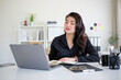 © wattana - Young professional woman in an elegant black blouse looks at her laptop while taking notes, portraying productivity and efficiency in a well-designed office.