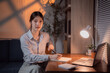 © Tj - Young Asian businesswoman working late at night in a cozy home office, taking notes on a notepad while focused on her laptop, surrounded by a warm lamp glow and plants
