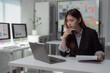 © Tj - Young Asian businesswoman holding a pen and contemplating while working on a laptop, analyzing financial data at an office desk, surrounded by documents and technology