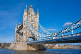 Tower Bridge stands majestically over the River Thames in London, showcasing its intricate architecture and vibrant blue cables against a bright sky, with people walking across.