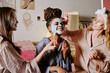 © AnnaStills - Medium shot of African American young woman with bun of dreadlocks wrinkling face while her biracial friends applying clay mask on her face
