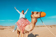 © EdNurg - Smiling female tourist riding a dromedary camel in the desert during a sunny day