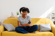 © Xavier Lorenzo - Happy young black woman with headphones using mobile phone relaxing on cozy sofa at home. Technology lifestyle concept
