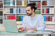 © Valerii Honcharuk - Young male teacher working in library sitting at desk with laptop books