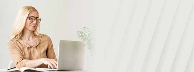 Aufkleber - A woman with long blonde hair and glasses is focused on her laptop while sitting at a stylish desk. Natural light fills the room, enhancing the cheerful atmosphere of her workspace, copy space