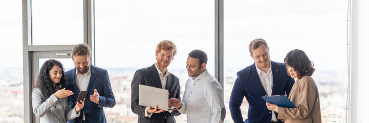 Aufkleber - A group of business professionals are having a meeting in a modern office. They are standing around a large wooden table, looking at laptops and tablets, and discussing work.