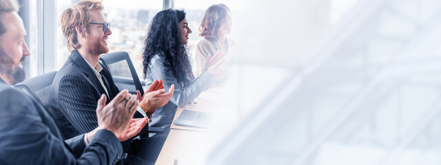 Poster - A group of business professionals sit at a table in a modern office, clapping their hands in appreciation as they listen to a presentation. They are dressed in formal attire