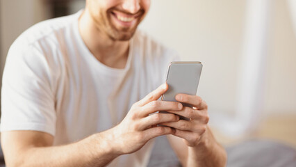Aufkleber - Unrecognizable Smiling Man Using Smartphone Mobile App Sitting In Bedroom At Home, Focus On Phone. Guy Texting On Cellphone With Social Media Application Concept. Cropped, Selective Focus, Panorama