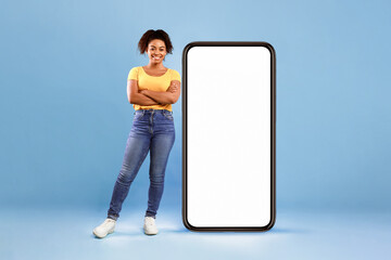 Poster - Beautiful African American lady standing near huge smartphone with empty white screen on blue studio background, space for mockup. Newest mobile application, website, advertisement concept