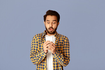 Poster - Handsome Indian man looking at his smartphone in shock over lilac background