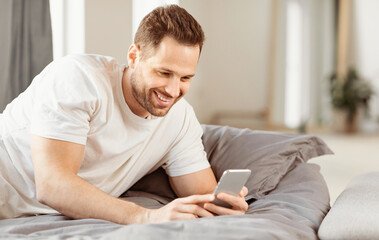 Aufkleber - Mobile App. Cheerful Guy Using Smartphone Lying In Bed In Bedroom At Home. Man Browsing Internet On Phone. Application For Cellphone Advertisement. Free Space For Text