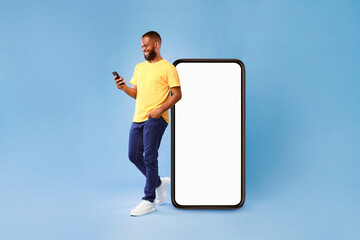 Poster - Handsome young African American man using mobile phone while standing near huge smartphone with empty screen over blue studio background. Mockup for website or application, space for advertisement