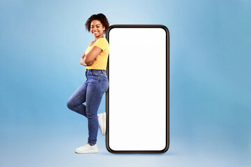 Poster - Full length of joyful African American woman leaning on big cellphone with empty space for mobile app on white screen, blue studio background. Smartphone display mockup, website or ad template
