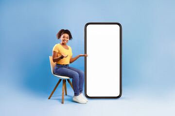 Poster - Full length of pretty African American lady sitting on chair next to big blank smartphone with mockup for mobile app or website design, blue studio background. Space for your online advertisement