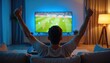 © Viktor - Man watching football game on TV at home. He celebrates scoring goal or team victory by raising arms, shows thumbs up. Emotional fan enjoys leisure time.