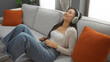 © Krakenimages.com - Woman relaxing on a couch with headphones, smiling, enjoying music while wearing casual clothing in a cozy living room with orange pillows and indoor plants