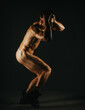 © qunica.com - A nude male model crouches in a dynamic pose, wearing boots, against a dark background.