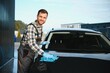 © Serhii - Car detailing, wash and cleaning concept. Young bearded male professional car wash worker in black rubber gloves, holding microfiber and wiping body of luxury car
