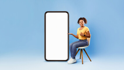 Poster - Cheerful young African American woman showing huge modern cellphone with blank screen, sitting on chair, offering newest mobile app over blue studio background, mockup