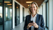 © Netaya - Happy middle aged businesswoman holds mobile cell phone uses cellphone in office. Smiling mature professional lady business owner entrepreneur uses smartphone while standing in hallway. concept as Hap