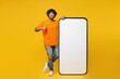 © ViDi Studio - Young smiling happy Indian man he wear orange t-shirt casual clothes big huge blank screen mobile cell phone smartphone with area show thumb up isolated on plain yellow background. Lifestyle concept.