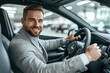 © ffunn - Portrait of a handsome man sitting in a new car at an auto showroom, smiling and holding the steering wheel while driving the vehicle with his hands on the wheel