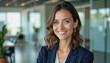 © f_bossa - A portrait headshot photo of a friendly professional CEO executive business worker: A smiling businesswoman with brown hair and light eyes confidently poses in a modern office lobby with glass wa