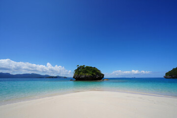 Poster - a tranquil beach scene with a small island in the background, featuring clear blue water and a bright blue sky. The sand is white and appears soft.