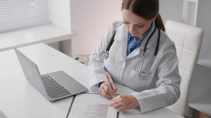 Poster - A dedicated healthcare professional in a white coat is focused on writing notes at a desk while an open laptop nearby shows their commitment to diligent patient care and recordkeeping
