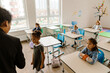 © Drobot Dean - A multiethnic group of students sits attentively at their desks in a bright, modern classroom. A girl walks toward the teacher while others observe, participating in an interactive learning activity.