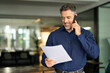 © insta_photos - Smiling middle aged senior professional businessman executive looking at documents talking on cell phone standing in office. Happy busy mature older business man holding papers making call at work.