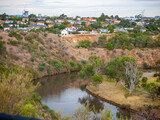 The Maribyrnong River winds through a suburban landscape in Melbourne, Australia, bordered by native trees, grasslands, residential houses, and sloping terrain along the riverbank.