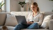 © M - Happy young woman works on laptop from home sitting on a couch.