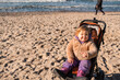 © Anton Dios - A cheerful toddler in a cozy sweater enjoys a sunny beach day sitting in a stroller on soft sand while gentle waves roll in nearby The sky is clear and blue