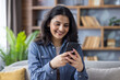 © Tetiana - Close-up photo of a smiling young Muslim woman sitting on the couch at home, holding a phone and looking at the screen