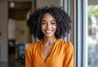 © Easy - A confident African American woman with curly hair wears an orange blouse while smiling at a modern office. Sunlight fills the space, creating a warm and welcoming atmosphere