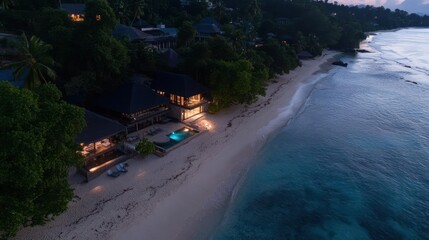 Poster - Aerial view of a tropical beach at dusk with luxury villas illuminated along the shoreline reflecting in calm ocean waters. Lush greenery surrounds the buildings