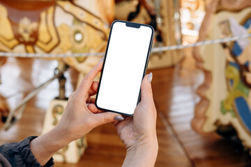 Mockup woman's hand holding using smart phone with white blank screen POV on the background of a carousel with horses, mobile app design and advertising, online marketing