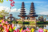 A serene landscape featuring traditional Balinese temples with tiered roofs, surrounded by vibrant flowers and a tranquil lake under a clear blue sky.