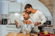 © bernardbodo - Two men happily embrace and smile while using a smart phone in a kitchen