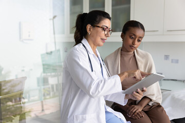 Positive mature Latin physician doctor showing checkup electronic result to young African patient woman sitting on couch in examination room, using digital tablet computer