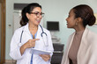 © fizkes - Positive friendly Latin female doctor talking to young Black patient woman, giving consultation, asking about healthcare, holding digital tablet for making notes in electronic records