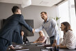 © fizkes - Two smiling male colleagues shake hands, closing profitable commercial deal at meeting with employees in office. Businessmen handshaking get acquainted, greeting partner, making agreement. Partnership