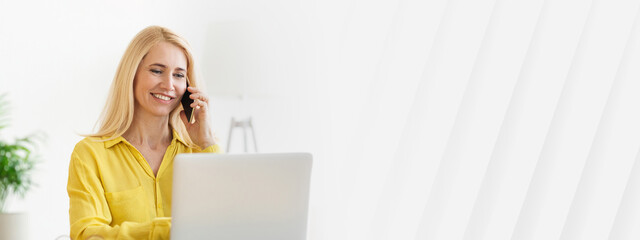 Poster - A woman with blonde hair wears a yellow shirt while talking on her phone and typing on a laptop at her home office. Natural light brightens the space, creating a cheerful atmosphere, copy space