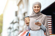 © Prostock-studio - Cheerful muslim girl with shopping bags and cup of coffee using smartphone, spending weekend at shopping mall