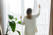© shurkin_son - Rare view indoor image of aged woman wife in white warm cardigan opening big window or curtains to let day light into the living-room with houseplants, enjoying weekend staying at home