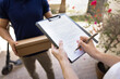 © fizkes - Professional doorstep delivery. Close up cropped view female recipient, delivery services client holds clipboard, put signature on confirmation form, while male courier stands with parcel box on porch